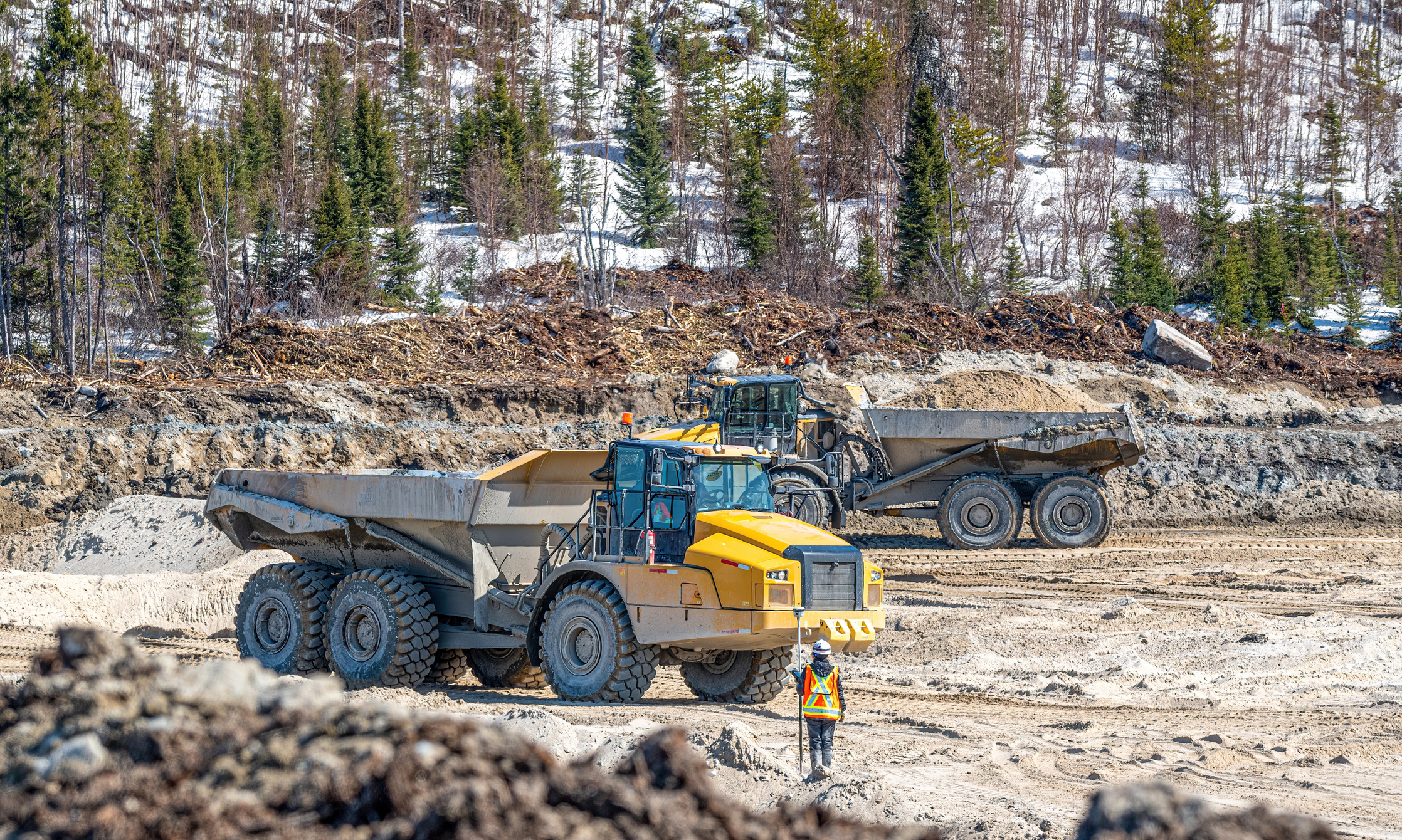 Two dump trucks transporting sand to a construction site, with a surveyor carrying out a survey using Heavy Equipment in British Columbia Wilderness